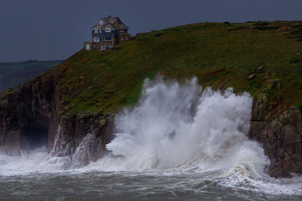 A massive wave breaks against a granite cliff. A house is perched precariously on the edge of the cliff